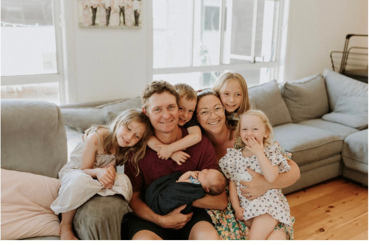 "Briony and the family crowded together and smiling for a photo sitting on the floor in front of a couch at their family home."