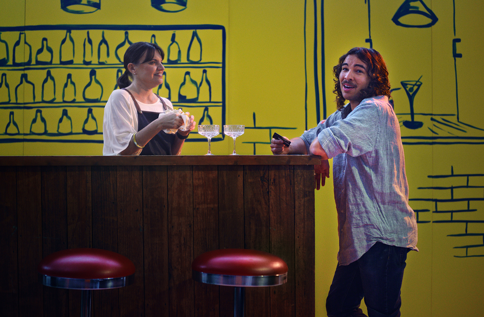 A woman in a white t-shirt with a denim apron stands behind a wooden bar, polishing a wine glass with two other glasses in front of her. A brunette man in a blue shirt and jeans is facing the camera in mid-conversation to the audience, holding a credit card. They are in front of a yellow and navy painted bar scene.