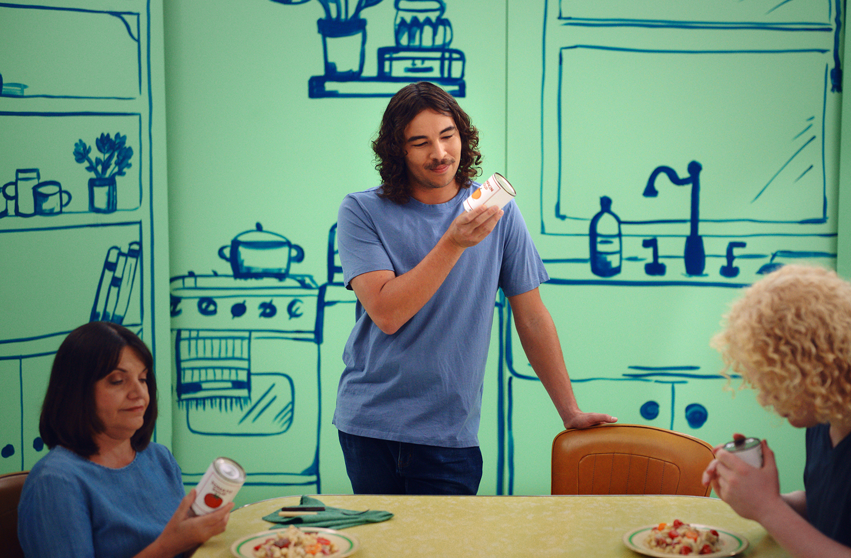 A man in a blue t-shirt is standing up at a kitchen table, holding a can up close to his face to read the packaging. Sitting at the table with him is a brunette lady and a man with curly blonde hair, also trying to read their own cans. They are in front of a green and navy painted kitchen scene.