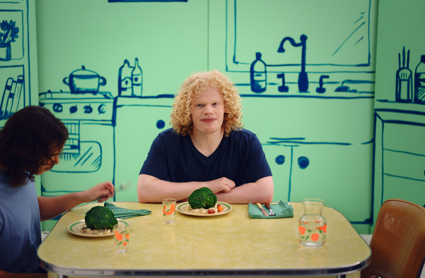 Two young men sit at a kitchen table with plates of broccoli pasta in front of them. A blonde man in a navy t-shirt is looking directly at camera with his arms folded on the table. A brunette man in a blue t-shirt is looking down at his plate with a fork in his hand. They are in front of a green and navy painted kitchen scene.