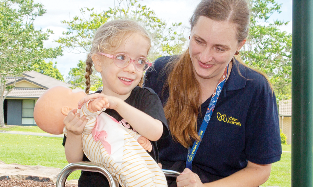 The young girl is holding a baby doll and in conversation with Vision Australia's occupational therapist at a playground