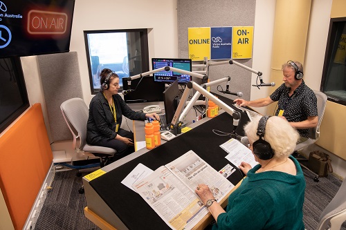 Three people sit in a radio studio and read newspapers over the air