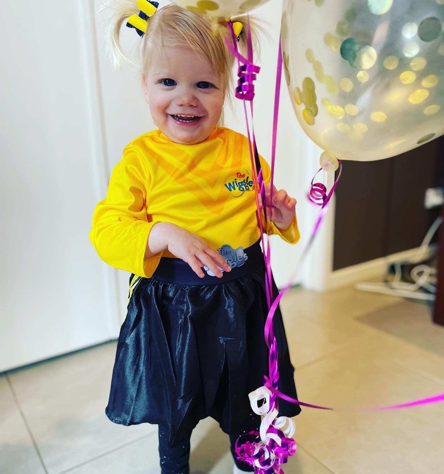 "Lucy wearing a yellow Wiggles shirt and skirt and smiling while holding a string attached to a helium-filled balloon."