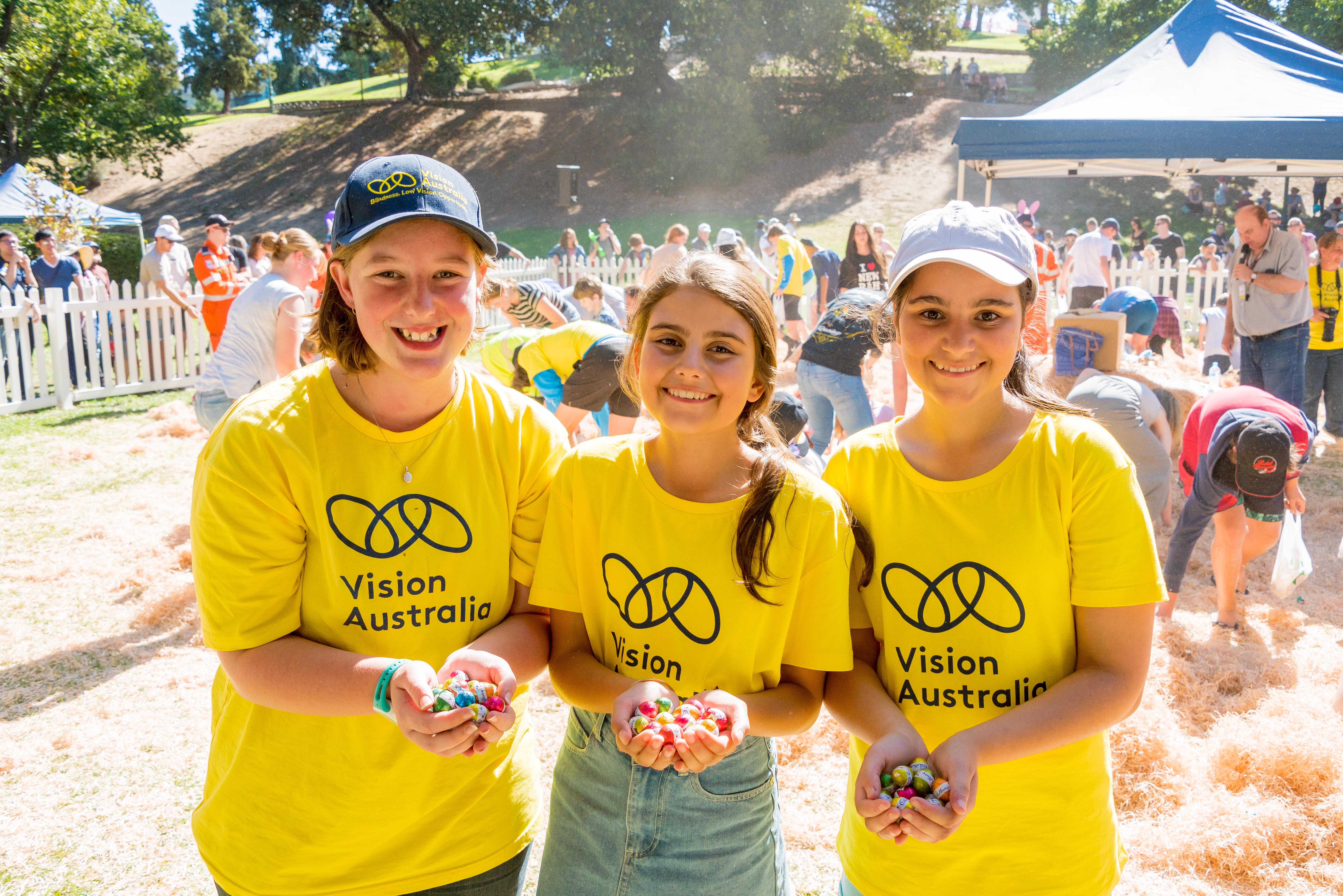 "Three volunteers in Vision Australia branded t-shirts each holding a handful of Easter eggs"