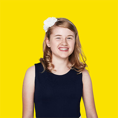 A young girl with blonde hair and wearing a black top smiles at the camera. The background is a bright yellow.
