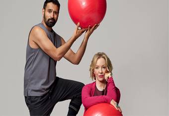 Rebecca Gibney is seated on the ground, resting her elbow on a red balloon. A man is standing above her, holding a red balloon over her head.