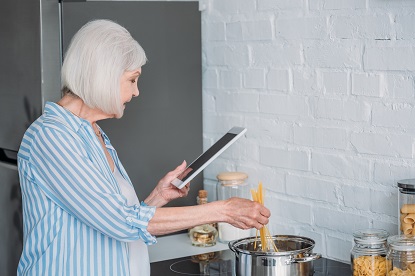 Image shows an older women cooking at a stove and reading from a tablet device