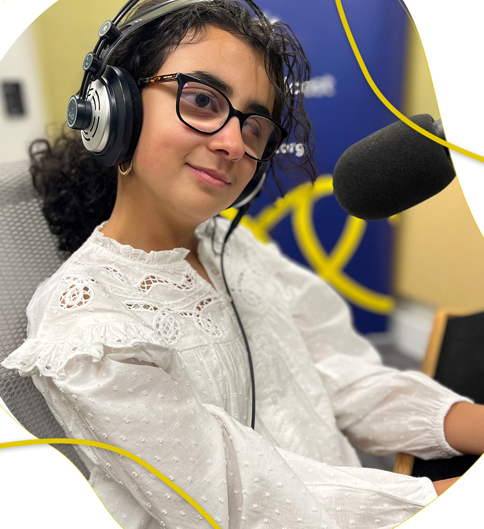 Haya, a girl with dark curly hair, is sitting in front of a studio microphone and wearing headphones, glasses and a white long-sleeved top.