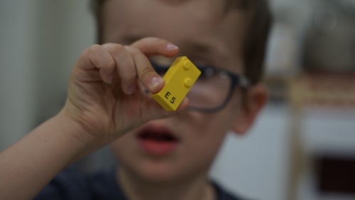 A young child holds a yellow LEGO Braille brick with raised bumps that correspond to the letter E