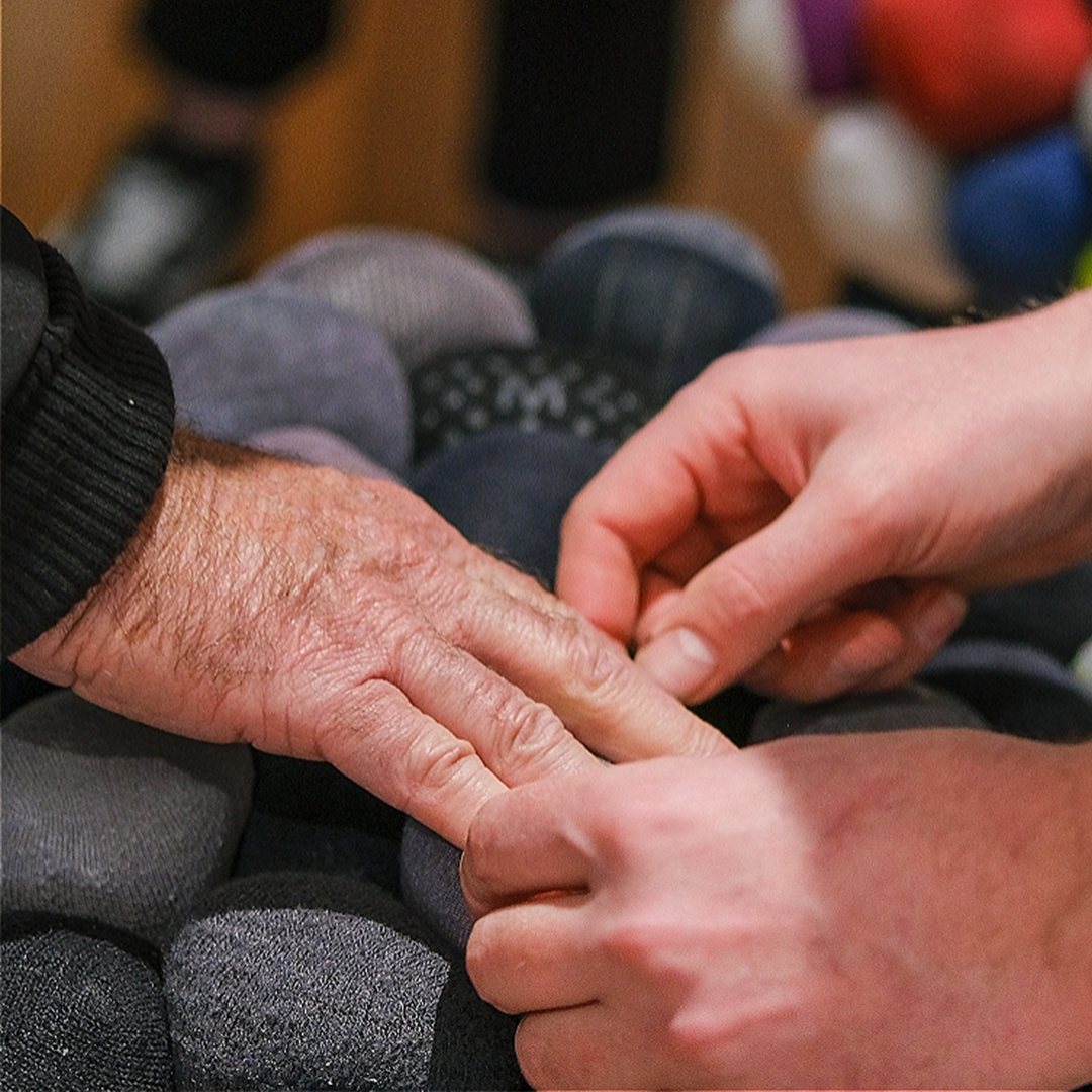 "A close-up view of a pair of hands guiding another person's right hand to touch the surface of a stool. The stool is made of multiple rolled-up pairs of old socks in various tones of grey. "