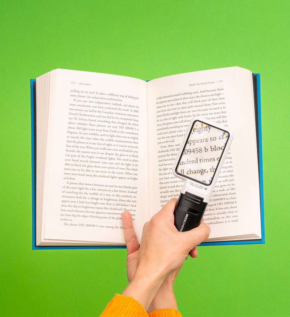 A person holds up a book on a bright green background, using a magnifier to make the book’s text bigger.