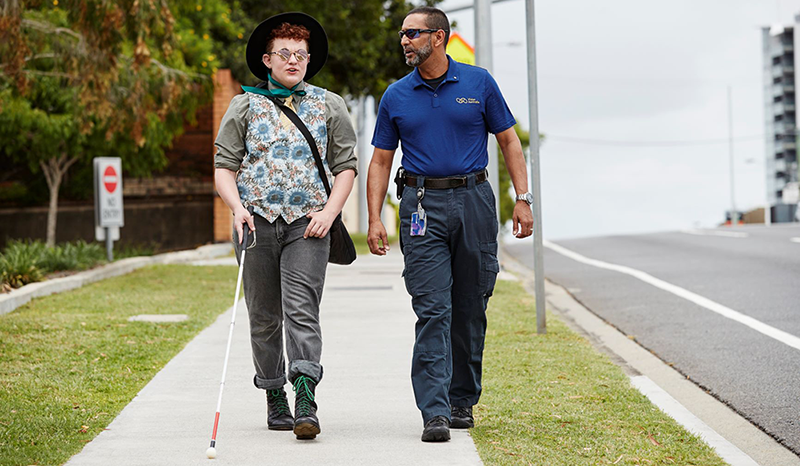 A Vision Australia Orientation and Mobility specialist next to a person who is blind or has low vision, with a white cane, walking together on the foothpath