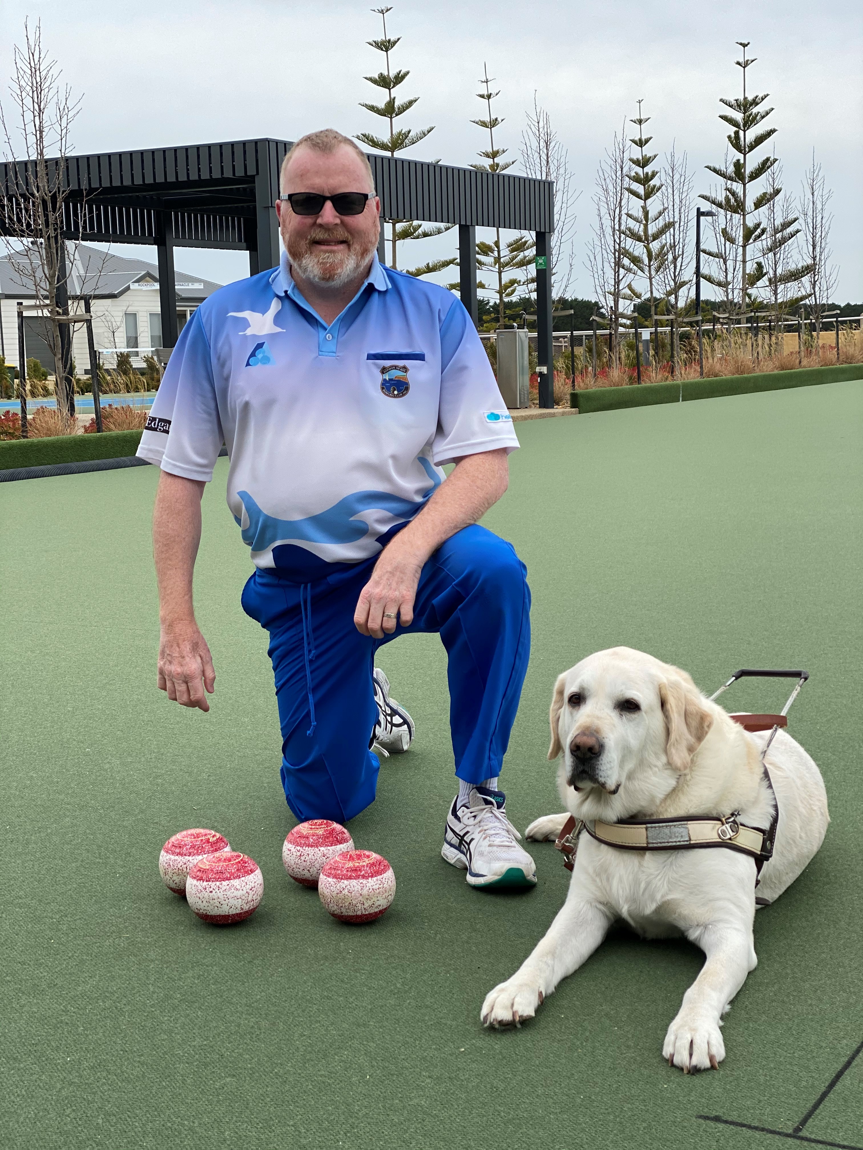 Greg Kennedy crouching with his dog guide Indrik on a Lawn Bowls court