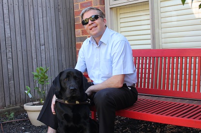 Image shows Hamish sitting outside on a red bench with his Black Labrador dog guide