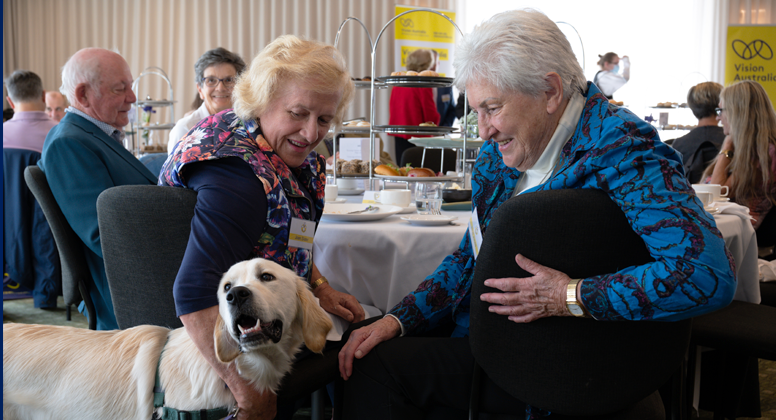 Two women are seated at a table and smiling whilst meeting a Seeing Eye Dog