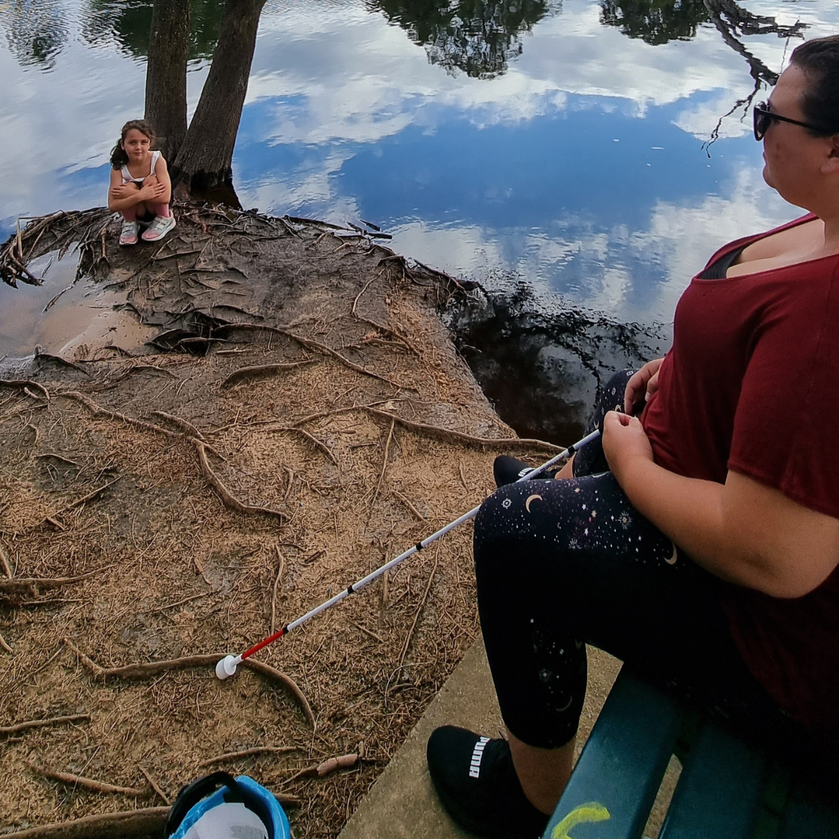 "Sarah sitting by a lake, white cane in hand, with one of her kids sat nearby leaning on a tree "
