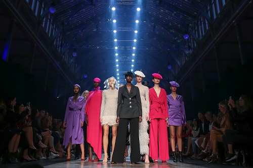 Seven fashion models stand together on a runway in a darkened room in front of a large crowd