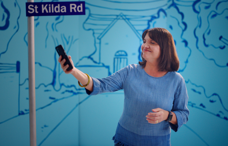 A woman wears a blue denim top, standing in front of a street sign in a light-and-dark blue handpainted street background. She holds in her hand to view the photo she took of the street sign.