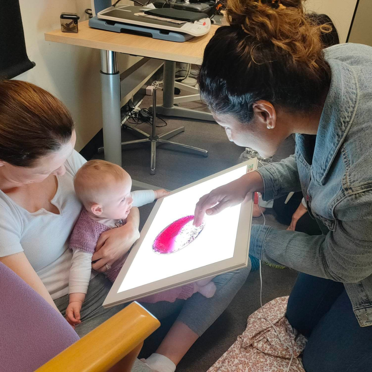 "Bonnie held by her mother as they look at a backlit screen held by a Vision Australia staff member who si holding a pink toy over it"