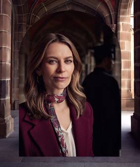 A woman with shoulder length brown hair, wide green eyes and a slight smile wears a burgundy jacket a white blouse and a silk patterned scarf. Behind her is the shadowy figure of a man. The picture of these people is overlayed against a building with sandstone block walls.