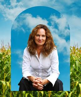 In the background a cornfield with floating white clouds in a bright blue sky. In the foreground a woman with shoulder length brown hair wearing a white shirt and black trousers sits with her elbows resting on her knees and her hands clasped.