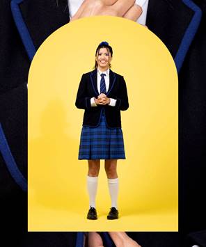 A teenage girl standing in a yellow archway with an anxious smile and her hands tightly clasped. She is dressed in a school uniform - white shirt and blue striped tie under a black blazer, a pleated, blue checked skirt, knee high white socks and black lace-up shoes.