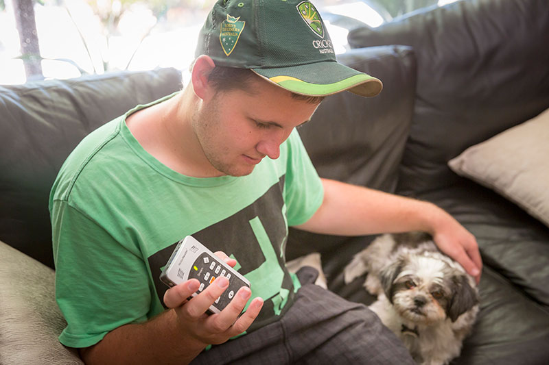 Zac is sitting on the couch, with his pet dog whilst listening and holding a handheld media player