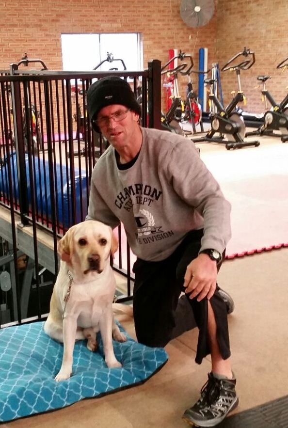 Grant is wearing gym clothes and is leaning down next to his Seeing Eye Dog in shot, with gym equipment in the background