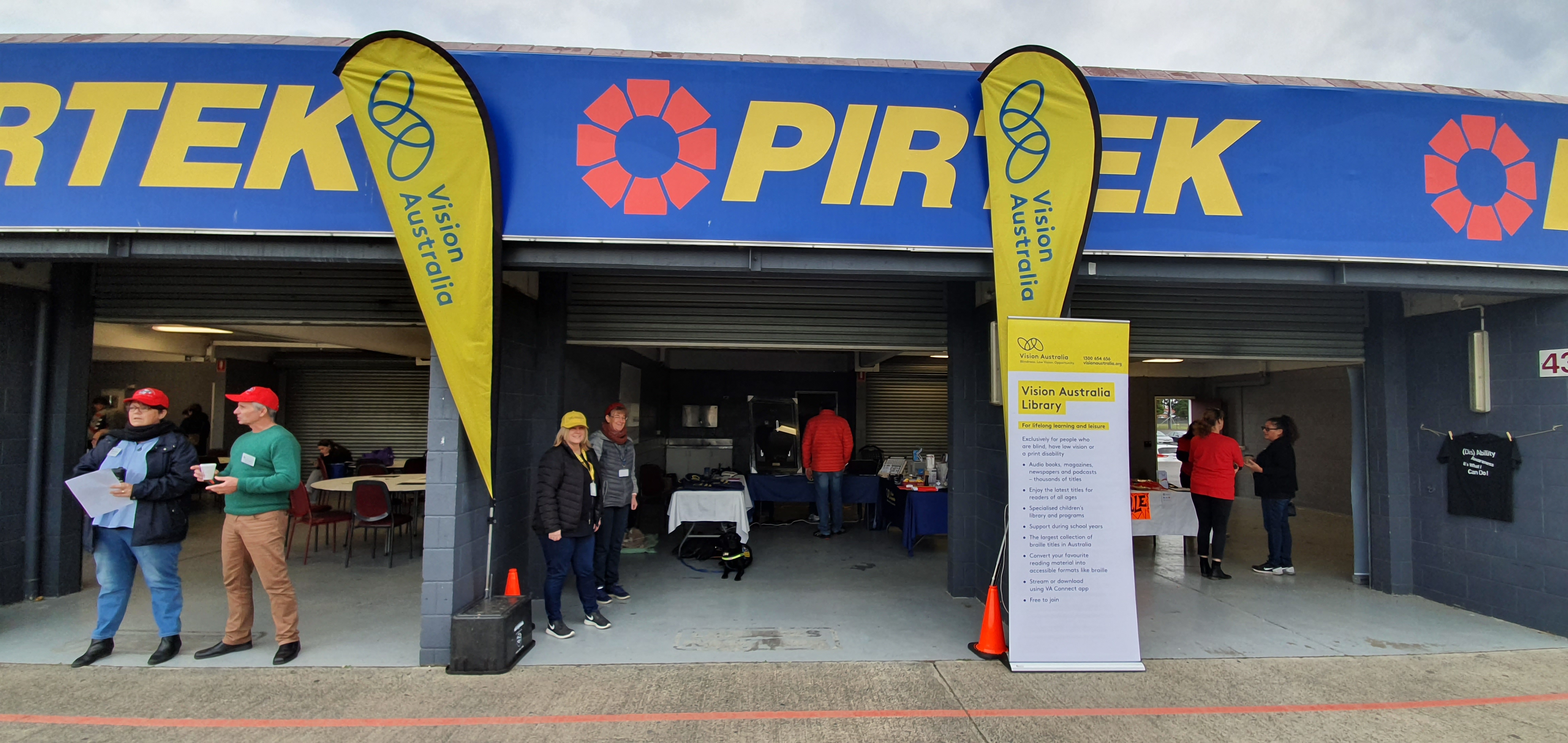 ""Vision Australia kiosk at In The Drivers Seat at Sandown Racecourse. Two yellow banners with the Vision Australia logo welcome people in.