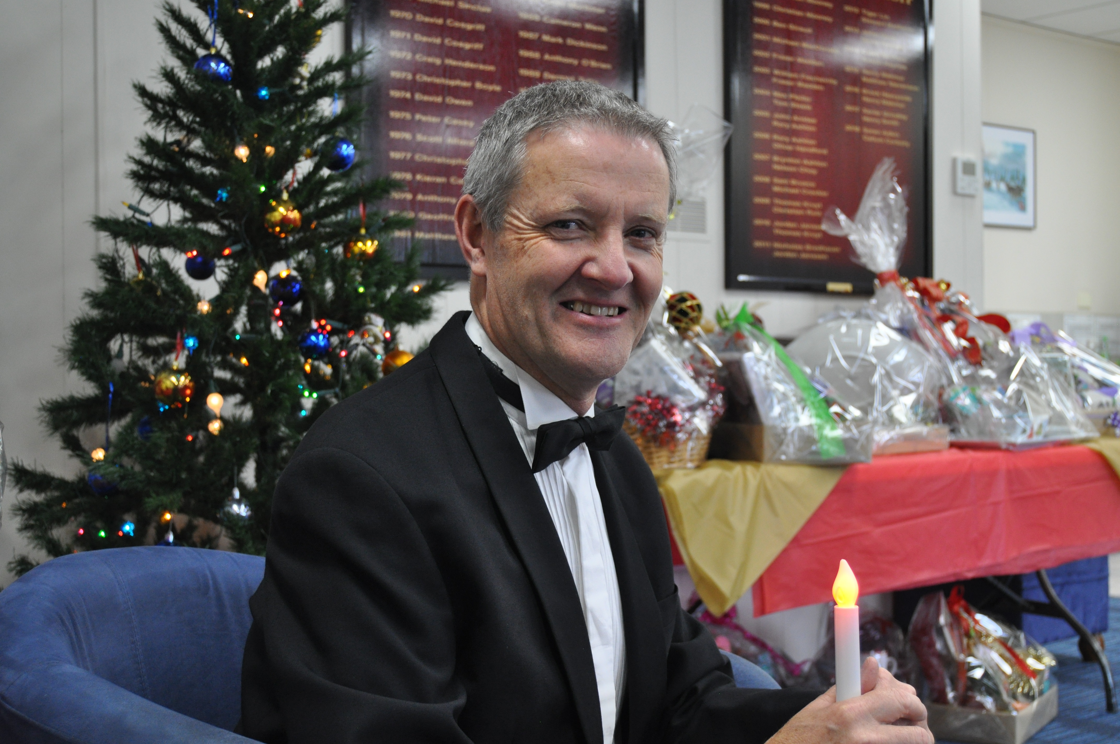 National Boys Choir co-artistic director, wearig a black dinner suit, holds a battery-powered candle with a Christmas tree and cellophane-wrapped gift baskets in the background.