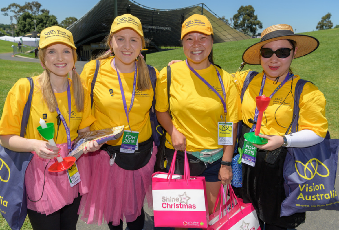 "Four volunteers from Carols by Candlelight stand at the Sidney Myer Music Bowl holding gift bags and smiling"