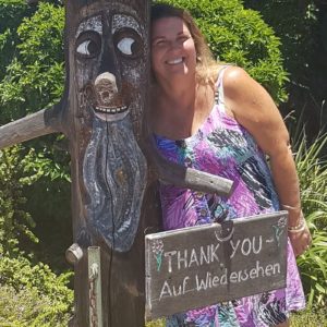 Teressa Andrews standing next to a smiling tree with the message "Thank you - Auf Wiedersehen" on a wooden sign in the foreground