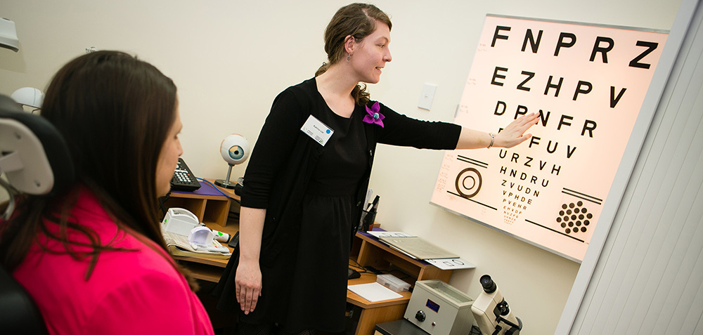 Eye specialist is pointing to a row on the eye chart in front of seated client.