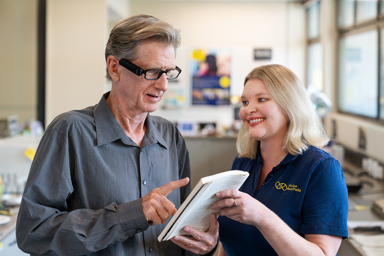 Phillip standing next to a Vision Australia staff member looking at a book and smiling