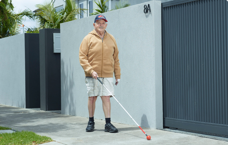 Man with a cane walks down a sunny street footpath