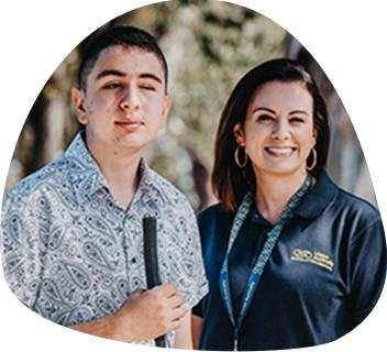 A Vision Australia staff member wearing a navy polo is standing beside a teenage boy who is holding a cane. They are both looking at the camera.