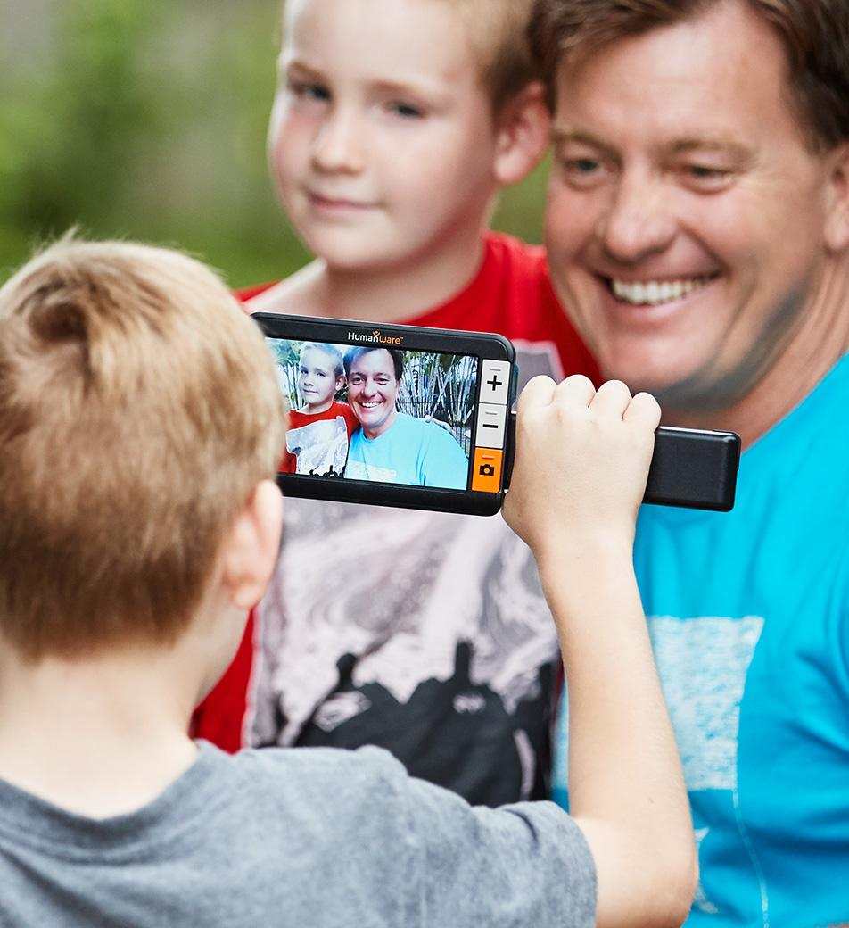 David poses with son as another son takes a photo using a handheld electronic magnifier device