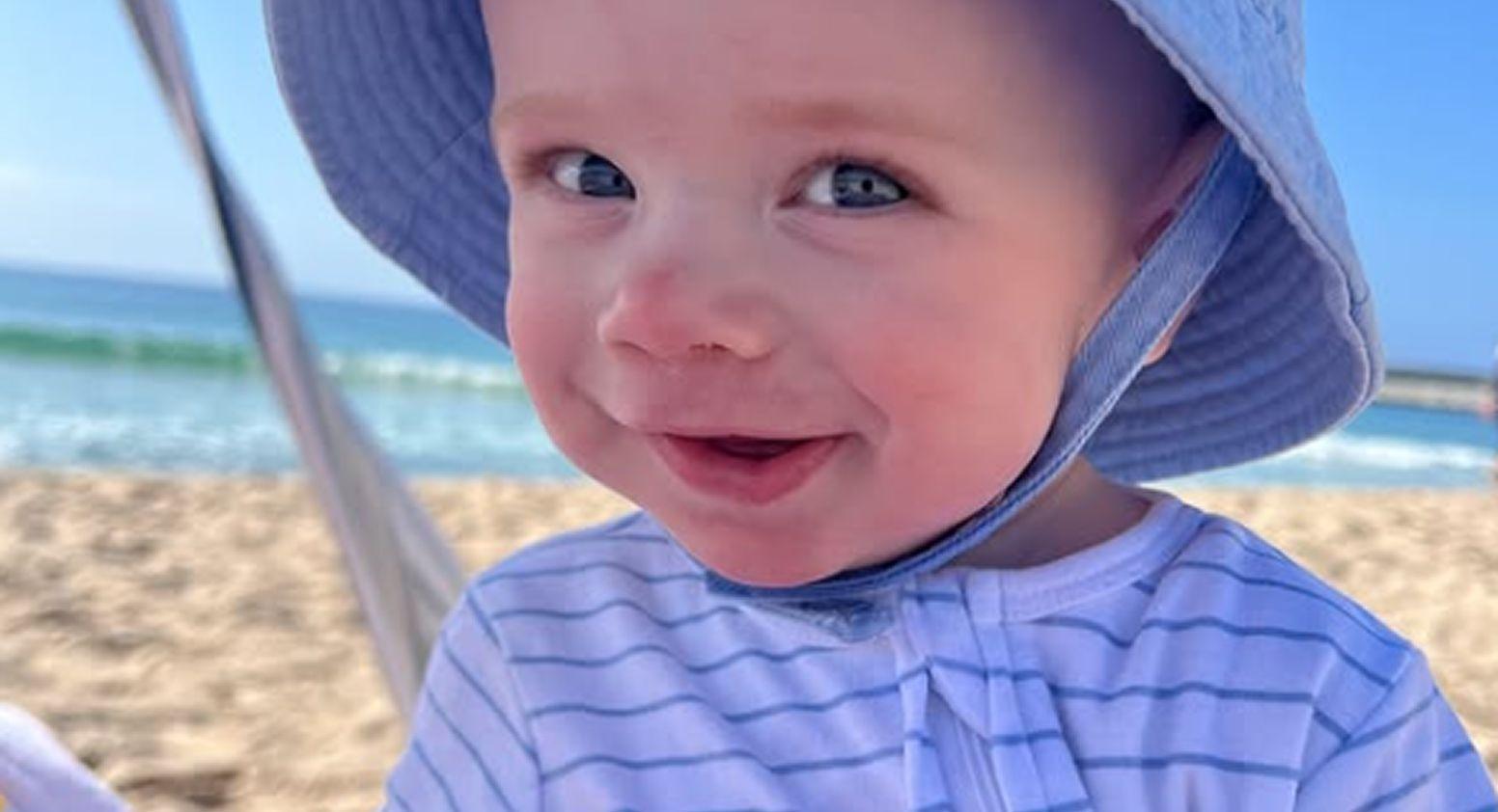 Flynn is on the sand at the beach, smiling to camera, and wearing a blue hat
