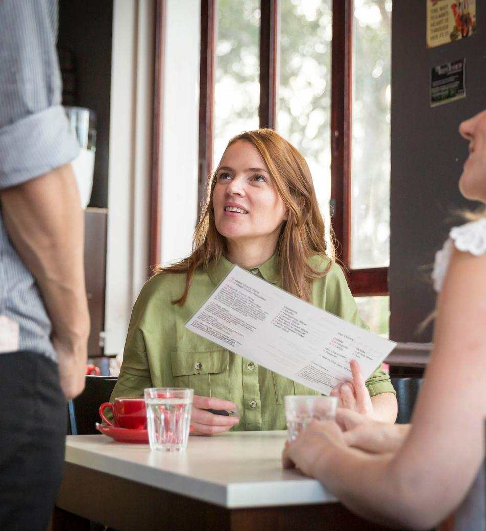 Two women sit across from each other at a cafe, smiling as they place an order with the server.