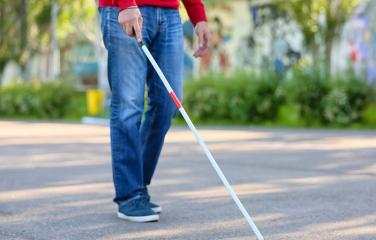 A man in jeans and a red jumper uses a cane to navigate on a footpath.