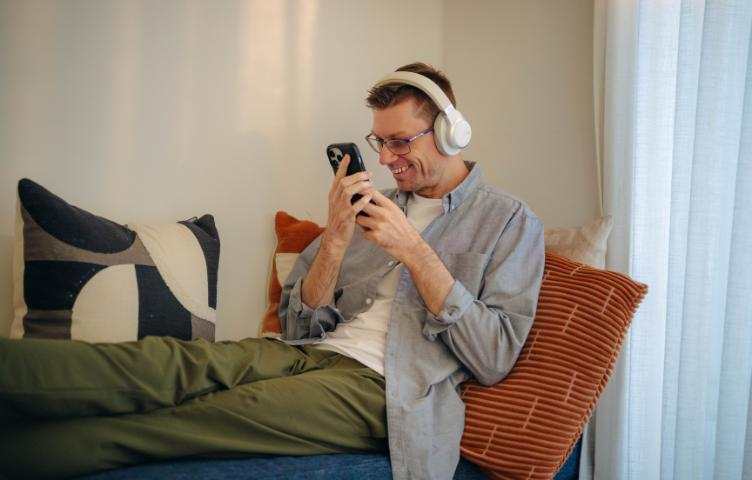 A man sits on his couch with headphones on, looking at this phone and smiling.