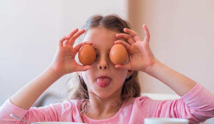 A young girl in a pink t-shirt pokes her tongue out and holds eggs over her eyes