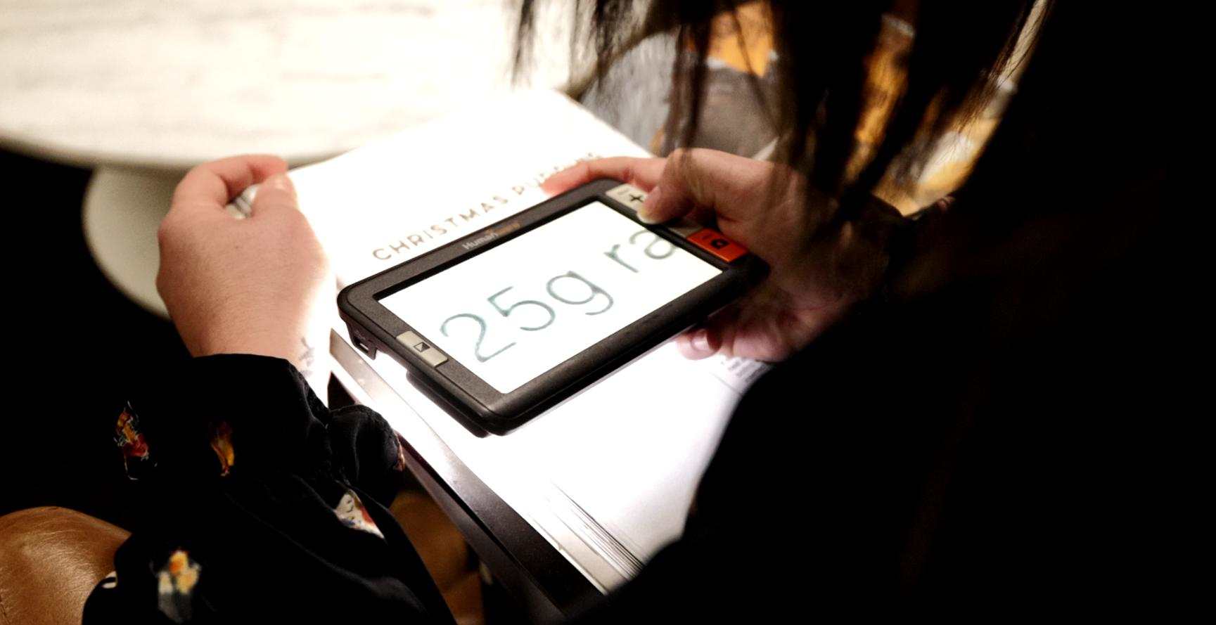 Person using a hand-held electronic magnifier device over a piece of paper. The text on paper is magnified on the hand-held device.